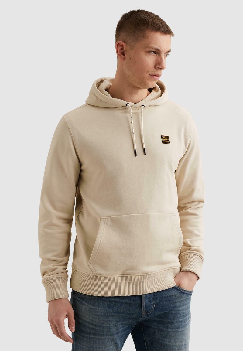 Jeune homme tourné vers la droite, portant un sweat à capuche beige avec une poche avant et un petit patch logo, associé à un jean bleu, mains détendues.