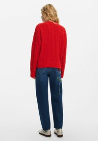 Maglione rosso a trama treccia con scollatura rotonda, abbinato a jeans blu scuro che presentano ricami floreali verdi sulla tasca posteriore.