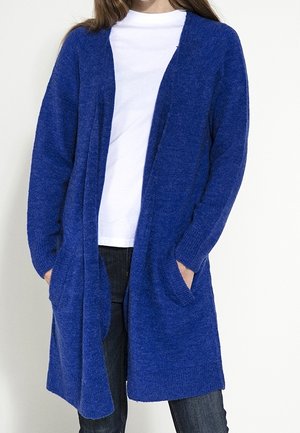 Person trägt eine lange blaue offene Strickjacke mit Taschen, ein weißes T-Shirt und dunkle Jeans vor einfachem Hintergrund.