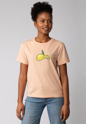 Pfirsichfarbendes Baumwoll-T-Shirt mit Rundhalsausschnitt und kurzen Ärmeln, das ein grafisches Motiv einer ganzen Zitrone und einer Zitronenscheibe mit grünen Blättern zeigt.