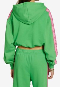 Groene cropped hoodie met roze accentstrepen langs de mouwen, voorzien van een capuchon en een zachte textuur. Gecombineerd met bijpassende groene broek.