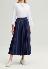 Gonna midi plissettata blu navy in tessuto testurizzato con vita alta elastica, abbinata a una maglietta bianca a maniche lunghe e sneaker bianche.