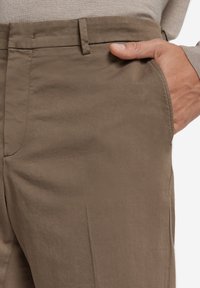 Braune Stoffhosen mit leichtem Glanz, ausgestattet mit einer Seitentasche und sauberen Nähdetails. Maßgeschneidert für eine figurnahe Passform.