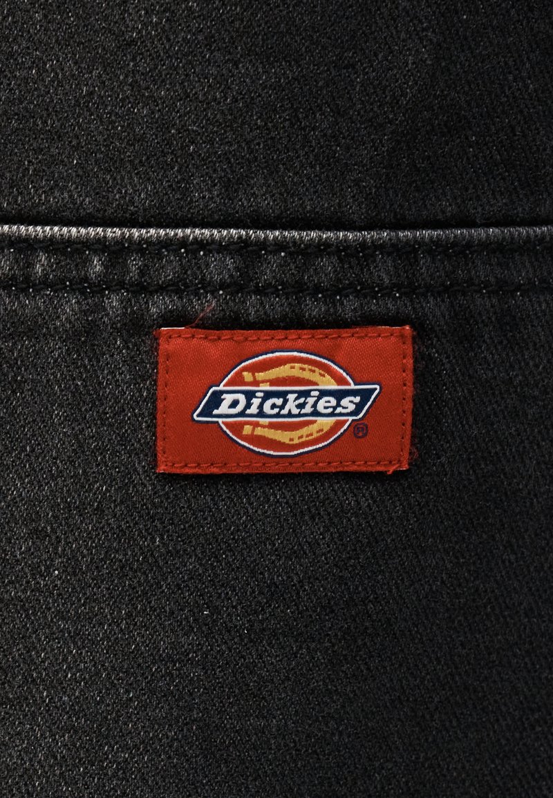 Rood rechthoekig stoffen label met het "Dickies"-logo, op zwarte spijkerstof genaaid nabij een naad.
