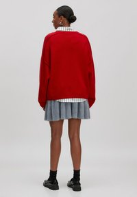 Roter übergroßer Pullover mit abfallenden Schultern, kombiniert mit einem hellgrauen, gefalteten Rock. Schwarze Schuhe und Knöchelsocken runden das Outfit ab.