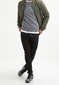 Person, der eine olivgrüne gesteppte Jacke, einen grauen Pullover über einem weißen Hemd, schwarze Hose und schwarz-graue Turnschuhe trägt, steht mit überkreuzten Beinen.