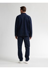 Mørkeblå denimjakke og bukser i en afslappet pasform. Har knapmanchetter og synlige syninger. Afsluttet med hvide sko.