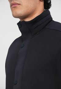 Veste navy avec un col haut, des boutons-pression et une texture lisse et soyeuse, arborant un design moderne et minimaliste.