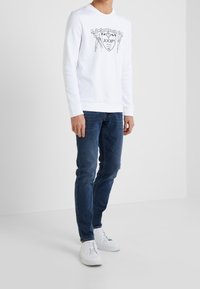 Sudadera blanca con un estampado gráfico que presenta figuras y texto en negrita. Combinada con jeans de mezclilla azul y zapatillas blancas. Diseño simple y casual.