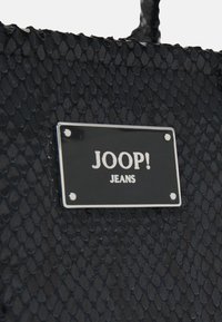 Musta teksturoidusta synteettisestä materiaalista valmistettu laukku, jossa on matelijakuviollinen pinta. Mukana hopeinen merkkilappu, jossa on kohokuvioitu "JOOP! JEANS".