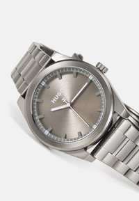 Montre en métal avec un cadran soleillé gris, des index argentés et un bracelet en acier inoxydable. Présente un boîtier rond et une couronne texturée.