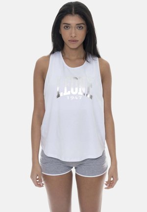 Femme aux longs cheveux foncés portant un haut blanc sans manches avec le texte "LEONE 1947" et un short de sport gris, debout devant un fond uni.