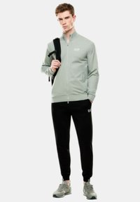 Giacca zip-up verde chiaro con colletto alto e tasche laterali, abbinata a pantaloni neri tapered. Scarpe da ginnastica grigie completano l'outfit.