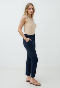 Canotta beige, pantaloni tapered navy con tasche laterali e flip flop con dettagli metallici. Tessuto liscio e design aderente.