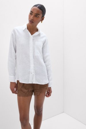 Frau trägt ein weißes langärmeliges Hemd mit Knöpfen und braune Shorts mit Spitzenbesatz und steht vor einem weißen Eckhintergrund.