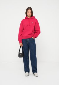 Rosa hettegenser med kengurulomme og snøring på hetten, kombinert med vide blå jeans. Tilbehøret er en liten svart håndveske med et logo.
