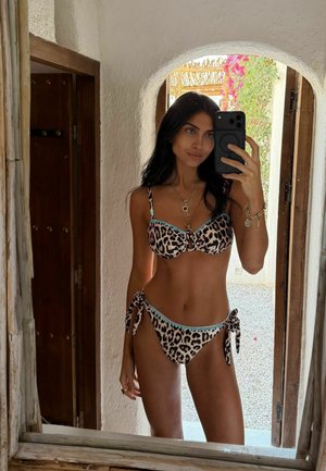 Donna con capelli lunghi e scuri che indossa un bikini stampato leopardato, scattando un selfie allo specchio in una stanza con un'apertura ad arco e piante all'aperto visibili.