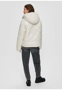 Puchowa, pikowana kurtka z dużym kapturem, z ukośnymi przeszyciami i luźnym krojem, noszona na ciemnoszarych dżinsach typu straight-leg i czarnych butach.