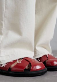 Punaiset nahkasandaalit kiiltävällä pinnalla, joissa on leveät ristiin menevät remmit ja musta kumipohja. Yhdistetty valkoisiin risareunaisiin housuihin.