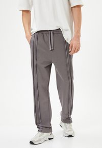 Pantalones de chándal grises en material suave, con franjas laterales, cintura elástica con cordón y puños ajustados. Combinados con zapatillas deportivas blancas.