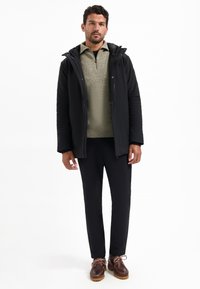 Man in zwarte capuchonjas, beige sweater met rits tot een kwart, zwarte broek en bruine leren schoenen, staande tegen een witte achtergrond.