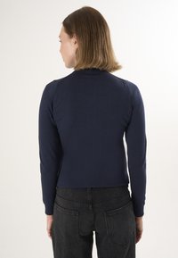 Granatowy, dopasowany top z długim rękawem, wykonany z teksturowanego materiału z prążkowanymi mankietami. Noszony z czarnymi, wysokim stanie dżinsami, widziany od tyłu.