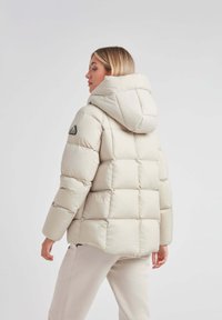 Beige pufferjakke med quiltet design, der har hætte og sort logo-patch på ærmet. Glat tekstur og afslappet pasform.