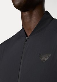 Schwarze Zip-Jacke mit hohem Kragen, aus strukturiertem Stoff und mit einem dezenten Logo auf der linken Seite. Glatter Reißverschluss und elegantes Design.