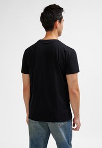 Man med kort mörkt hår som bär en svart t-shirt och blå jeans, stående med ryggen mot kameran framför en enkel vit bakgrund.