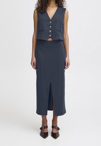 Nainen, joka on pukeutunut laivastonsiniseen hihattomaan nappivestiin ja siihen sopivaan korkeavyötäröiseen midi-hameeseen, jossa on etuhalkio, sekä tummanruskeisiin kärjellisiin korkokenkiin.