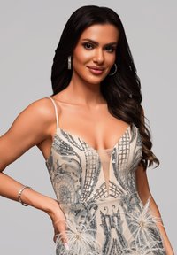 Robe beige avec des motifs complexes en sequins argentés, décolleté transparent et accents en plumes. Le mannequin porte des boucles d'oreilles créoles et un bracelet.