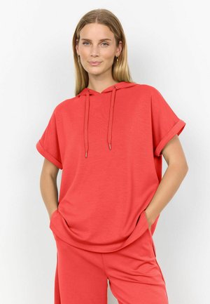 Kobieta o prostych blond włosach ubrana w koralową bluzę z krótkim rękawem i pasujące spodnie, stoi z rękami w kieszeniach na białym tle.