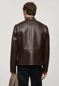 Veste en cuir marron au design ajusté, avec des détails de coutures visibles et des zips aux poignets. Texture lisse, silhouette structurée.