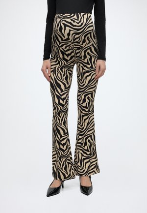 Žena u bež i crnim zvonolikim hlačama s uzorkom zebre, crnoj dugoj majici i crnim šiljastim visokim petama, stoji na bijeloj pozadini.