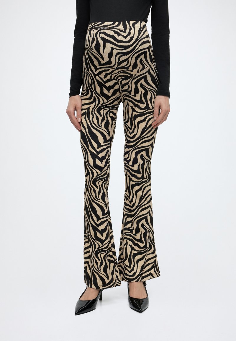 Mujer con pantalones acampanados estampados de cebra en beige y negro, camiseta negra de manga larga y tacones altos negros puntiagudos, posando contra un fondo blanco.