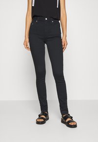 Jeans negros ajustados de cintura alta, con cinco bolsillos, cierre de botón metálico y cremallera, combinados con sandalias negras. Tela de mezclilla suave.