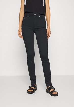 Jeans negros ajustados de cintura alta, con cinco bolsillos, cierre de botón metálico y cremallera, combinados con sandalias negras. Tela de mezclilla suave.