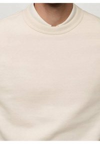 Mężczyzna ubrany w gładki, jasnobeżowy sweter z okrągłym dekoltem, widoczna tylko szyja i górna część klatki piersiowej.