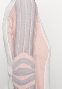 Vue latérale d'un pull en tricot présentant des blocs de couleur rose pastel, lavande et blanc avec des textures côtelées et rayées.
