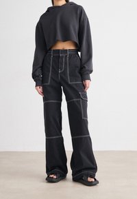 Zwarte cropped hoodie met lange mouwen, gecombineerd met brede zwarte broek met witte stiksel en een zijzak. Schoeisel is zwarte sandalen.