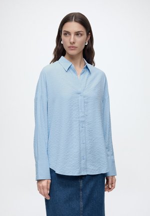 Femme aux longs cheveux bruns porte une chemise bleue claire texturée à boutons et une jupe en jean bleu foncé sur un fond blanc uni.