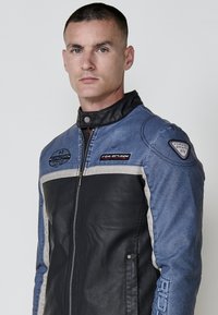 Plavi traper i crna ko�žna moto jakna sa srebrnim detaljima; ima zatvaranje na zip, visok ovratnik i brendirane zakrpe na ramenima.