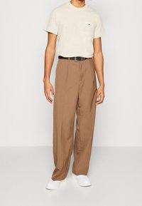 Beige bavlněné tričko s logem, hnědé široké kalhoty, černý pásek a bílé tenisky. Minimalistický design s hladkou texturou.