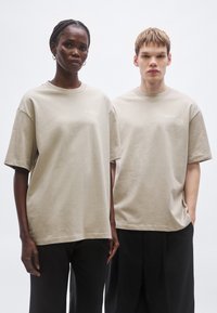 Tricouri beige supradimensionate din țesătură moale de bumbac, cu mâneci scurte și gât rotund. Detaliu subtil cu logo aproape de piept.