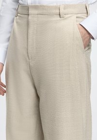 Homme portant un pantalon beige habillé avec la main droite dans la poche, manche de chemise blanche visible sur fond clair.