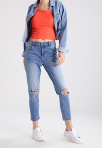 Jeans strappati blu in denim, top corto arancione e giacca di jeans in denim chiaro. Sneakers bianche completano il look casual.