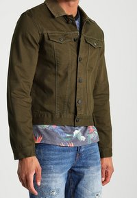 Chaqueta de mezclilla verde oliva con botones en el frente, dos bolsillos en el pecho y un cuello de pana marrón, combinada con una camisa de estampado tropical.