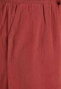 Tissu couleur rouille avec une surface texturée et froissée. Présente une taille élastique et des plis subtils pour plus de détails.