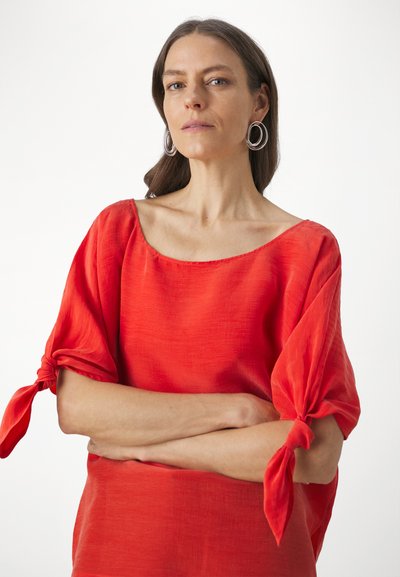 Red linen top with a wide, rounded neckline and short sleeves tied at the elbows. Smooth texture, lightweight fabric, and a relaxed fit.