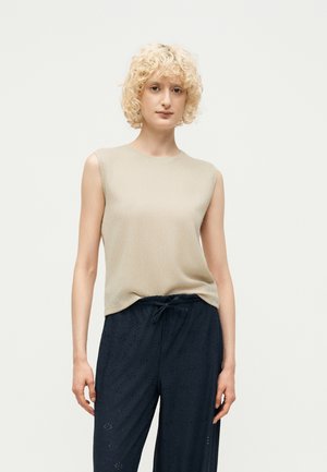 Femme aux cheveux blonds bouclés portant un haut beige sans manches et un pantalon noir ample avec des détails œillets, debout contre un fond uni.